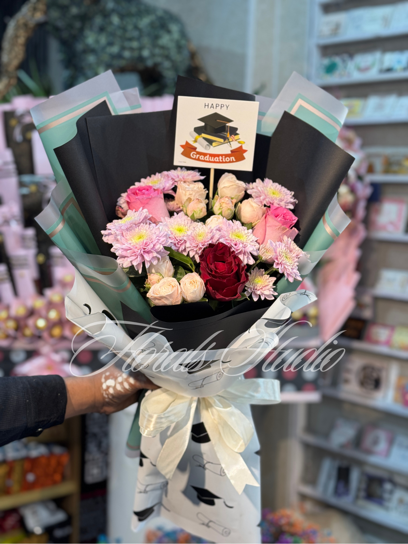 "Grad Bloom Elegance – Graduation bouquet with pink chrysanthemums, white roses, and red roses wrapped in black and white graduation-themed paper, available for fresh flower delivery in Lahore."