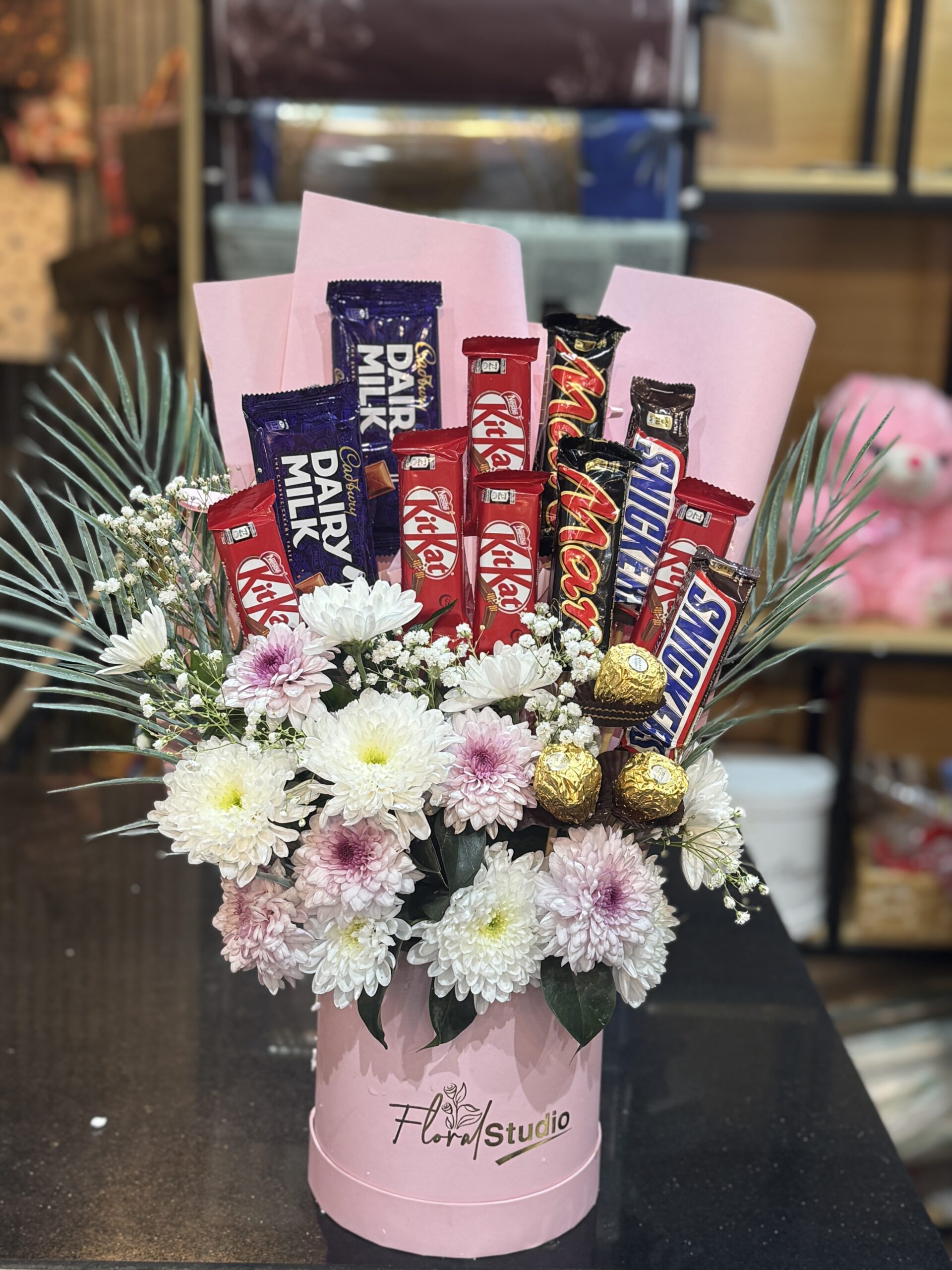 A luxury pink box with fresh white and pink chrysanthemums, baby's breath, and premium chocolates including KitKat, Snickers, Dairy Milk, and Ferrero Rocher. Perfect for gifting from a flower shop in Bahria Town, Lahore.