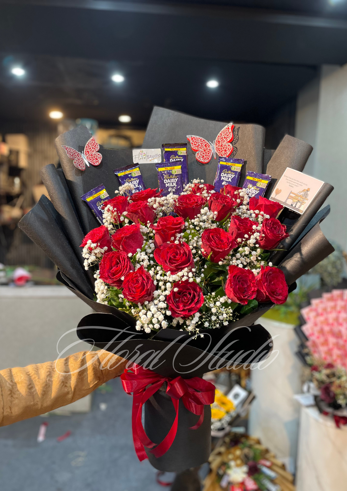 Sweet Scarlet Surprise bouquet featuring fresh red roses, white baby's breath, Cadbury Dairy Milk chocolates, elegant black wrapping with a red ribbon, and decorative butterfly accents, available for online flower delivery in Lahore.